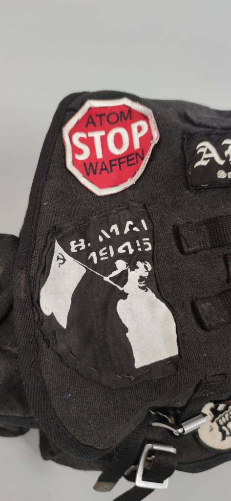 Detailaufnahme des Rucksacks. Zu sehen sind zwei Aufnäher, auf einem steht in weißer Schrift auf schwarzem Hintergrund "8. Mai 1945" und eine Figur schwenkt eine Flagge mit dem Hammer und Sichel-Symbol. Der zweite Aufnäher sieht aus wie ein rotes Stoppschild mit der Aufschrift "Atomwaffen Stop".t