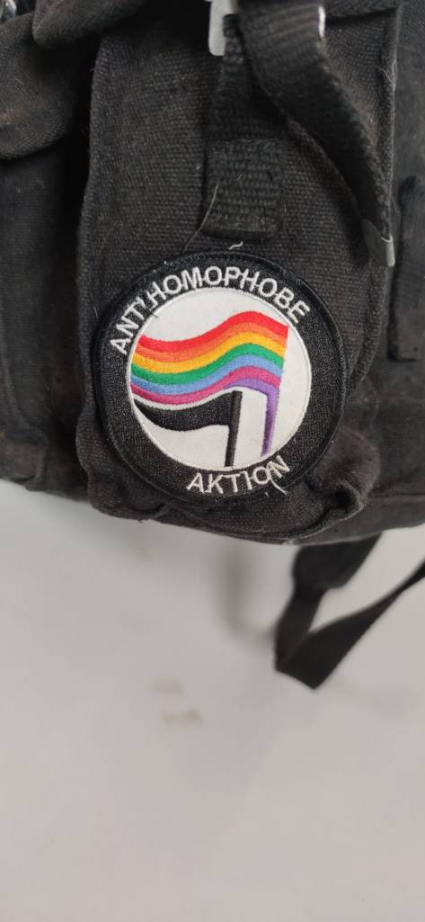 Detailaufnahme des Rucksacks. Der Fokus liegt auf einem runden Aufnäher mit schwarzer- und Regenbogenflagge. Außen am Kreis steht "Antihomophobe Aktion".