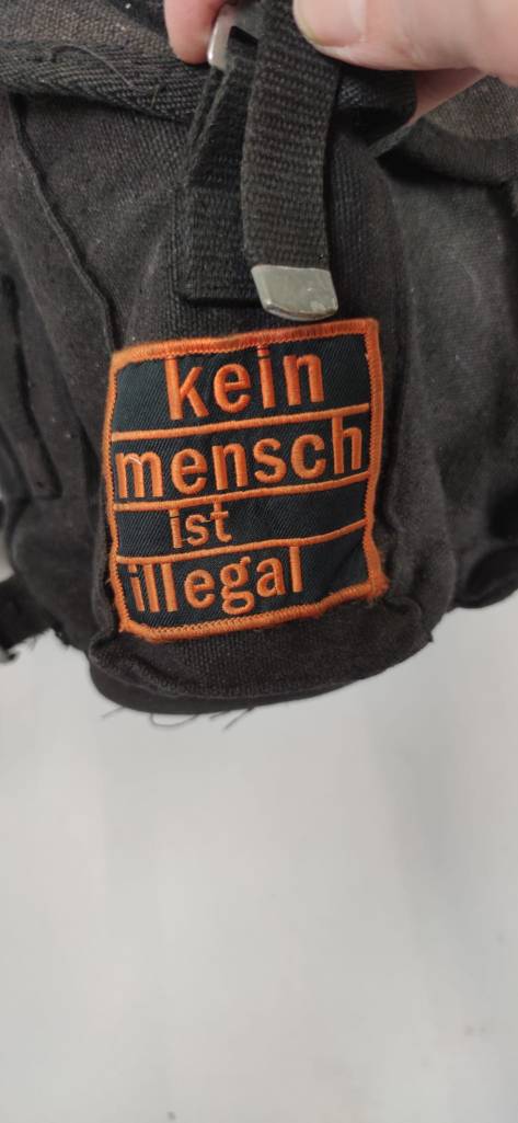 Detailaufnahme des Rucksacks. Fokussiert ist ein quadratischer Aufnäher mit der orangen Schrift "kein Mensch ist illegal". Oben im Bild sieht man eine Hand, die den Rucksack hält.