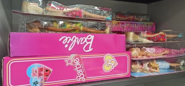 Das Bild zeigt einen Stapel Barbiepuppen in ihren Verpackungen. Sie liegen in einem Schrankfach.