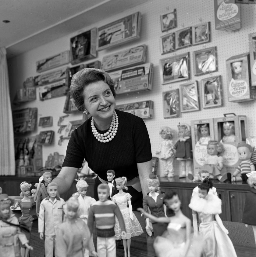 Auf dem schwarz-weiß Foto steht Ruth Handler vor einer Kollektion Barbiepuppen und lächelt. Sie trägt eine Kurzhaarfrisur und eine helle Perlenkette. Im Hintergrund sind an einer Wand weitere Kinderspielzeuge, wie Gewehre und Babypuppen, erkennbar.