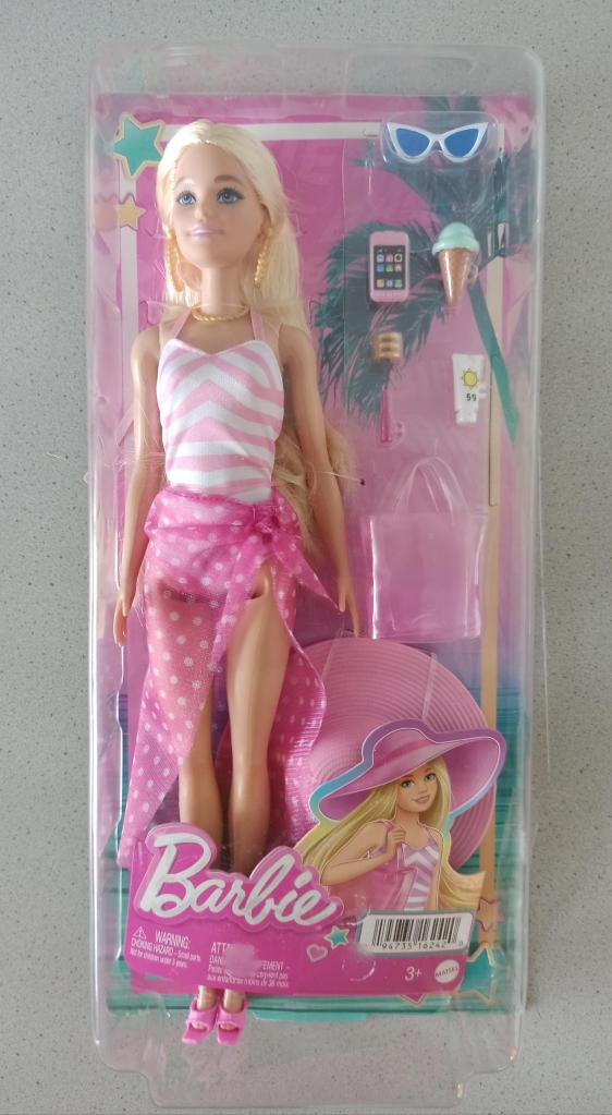 Das Foto zeigt eine blonde Barbie in einer größeren pinken Verpackung. Sie trägt einen rosafarbenen Badeanzug und ein pinkes Strandtuch mit weißen Punkten um die Hüfte. Neben der Puppe sind Accessoires aus Plastik in der Verpackung: eine Sonnenbrille, ein Handy, ein Eis, Sonnencreme, Schmuck, eine Tasche und ein Sonnenhut.