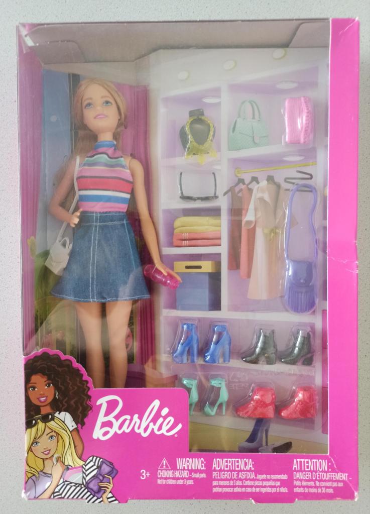 Dieses Foto zeigt eine blonde Barbiepuppe in einer größeren pinken Verpackung. Sie trägt einen Jeansrock und ein gestreiftes Top. Sie steht neben einem aufgemalten Kleiderschrank, in dem echte Schuhe und Taschen aus Plastik für die Puppe stehen.