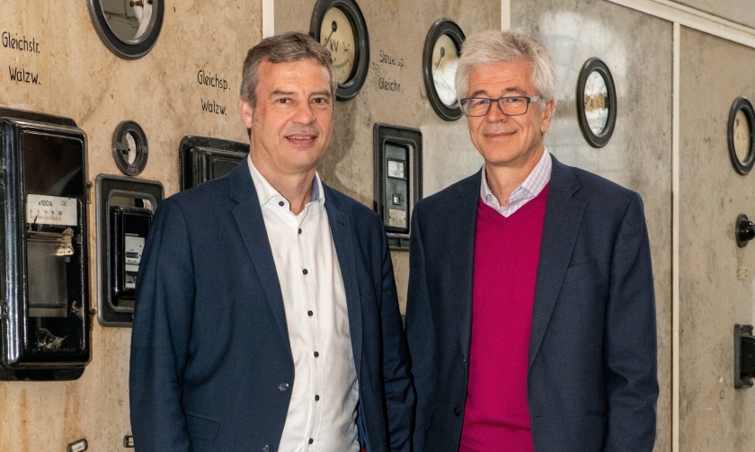 Auf dem Bild stehen Dietmar Osses und Walter Hauser vor einer Schaltwand in der ehemaligen Elektrozentrale der Zinkfabrik Altenberg und lächeln. Dietmar Osses steht links und trägt ein blaues Sakko, Walter Hauser steht rechts neben ihm und trägt einen pinken Pullover unter einem dunkelgrauen Sakko.