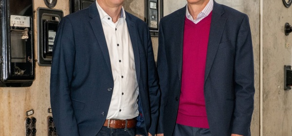 Auf dem Bild stehen Dietmar Osses und Walter Hauser vor einer Schaltwand in der ehemaligen Elektrozentrale der Zinkfabrik Altenberg und lächeln. Dietmar Osses steht links und trägt ein blaues Sakko, Walter Hauser steht rechts neben ihm und trägt einen pinken Pullover unter einem dunkelgrauen Sakko.