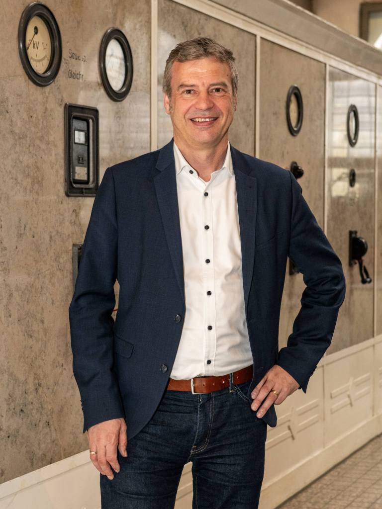 Auf dem Foto steht Dietmar Osses vor einer Schaltwand in der ehemaligen Elektrozentrale der Zinkfabrik Altenberg. Er trägt ein blaues Sakko, Jeans und ein weißes Hemd. Er lächelt in die Kamera.